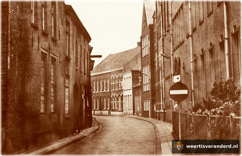 Vanaf de Korenmarkt kijken we de Schoolstraat in. Aan de linkerkant is de zijgevel van Hotel de Engel dichtgespijkerd, want het pand staat te wachten op restauratie. Rechts is de grote voorgevel van het complex Saint Louis. Op de plaats van