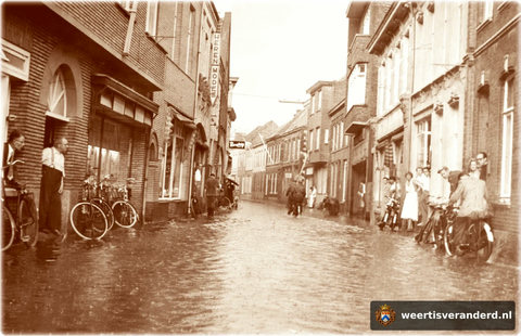 Wateroverlast kwam vaak voor in de Hoogstraat. Uiterst rechts de kapelanie met kapelaan Lemmens in de deur en naast hem huishoudster Klaartje van Riet. Links kijkt meester Vroemen uit het raam en staat Väös Janssen de schade te bekijken. Di