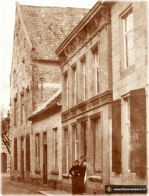 De fraai geklede dames staan in de deur opening van het pand van de familie Vaessen. In het huis ervoor met de luiken woonde de familie Caris. Het pand op de hoek heeft nog een hoge zijgevel.