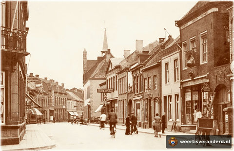Rechts zien we een typische transportfiets voor slagers en bakkers. Een beetje verder aan de rechterkant van de straat heeft Coöperatie Ons Belang inmiddels een uithangbord gekregen. In het hoge pand midden op de foto met het uitverkoop-bor