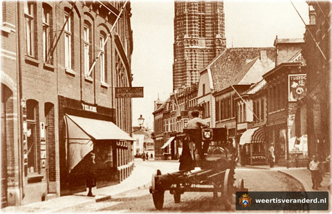 De lege bierwagen van de WERTHA, met “Frits vanne Wertha” op de bok, ratelt met zijn ijzeren velgen over de basaltkeien. In de zaak links, met de luifel, is lunchroom ’t Kelderke van de familie Geenen gevestigd.